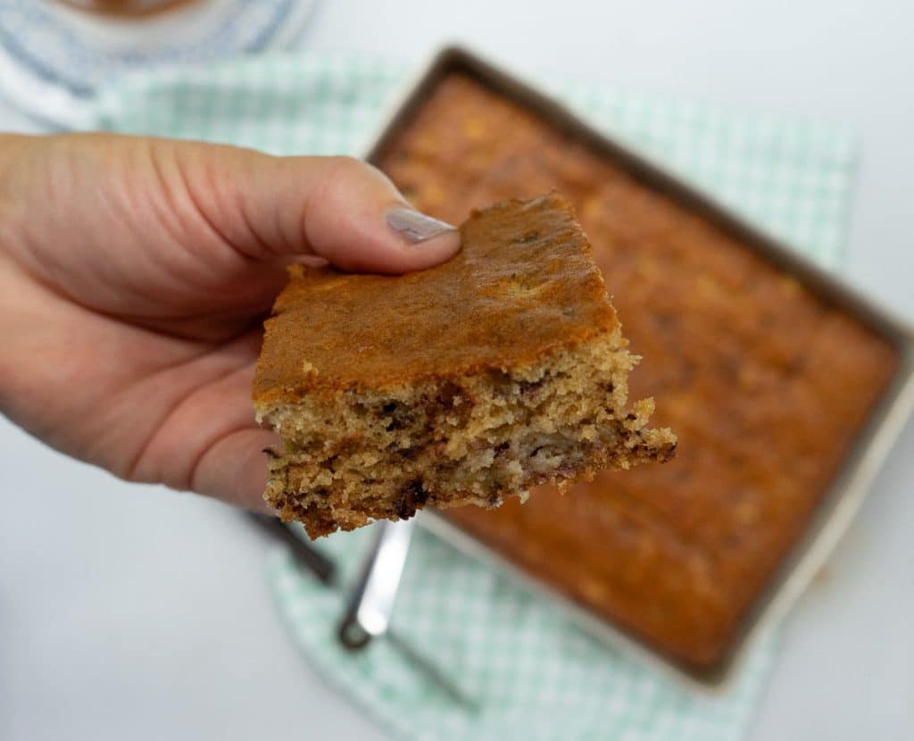 hand holding a piece of banana cake over the whole pan of cake