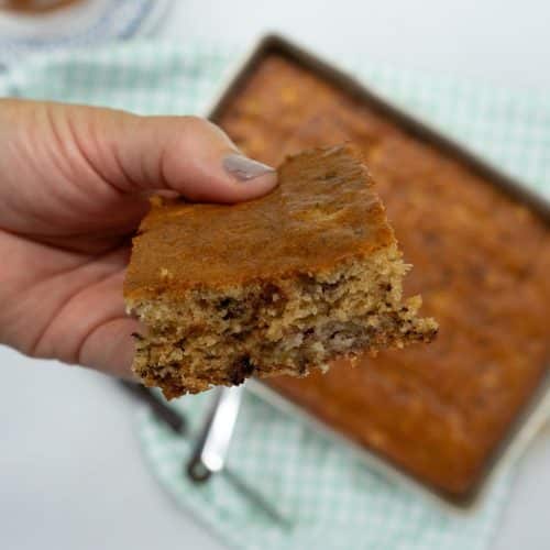 hand holding a piece of banana cake over the whole pan of cake