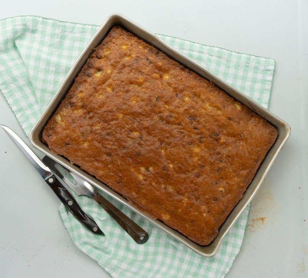 9x13 pan of banana snack cake with a knife and a spatula beside the pan, pan is sitting on a green and white gingham napkin on a white countertop