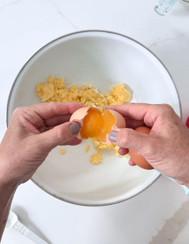 adding eggs to the butter and sugar combination in a white bowl, hands shown cracking egg