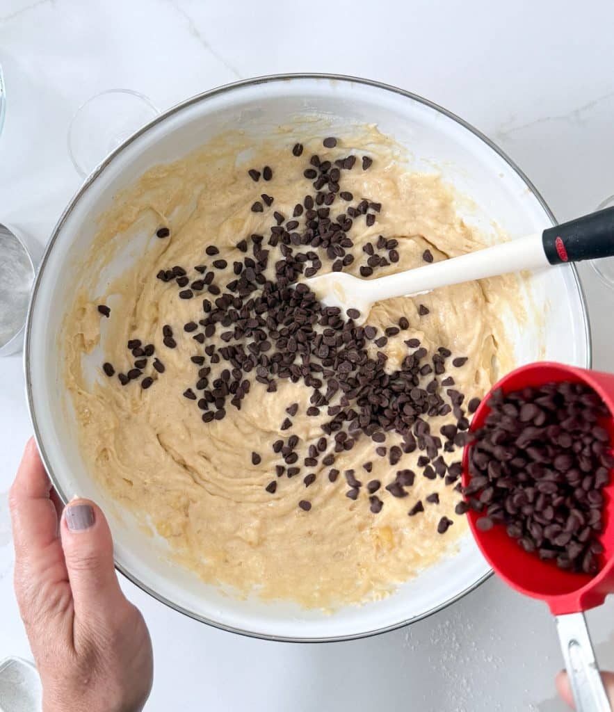 adding mini chocolate chips to the cake batter. hand holding bowl in lower left hand corner, chocolate chips falling out of a red measuring cup in right hand corner. spatula in the bowl