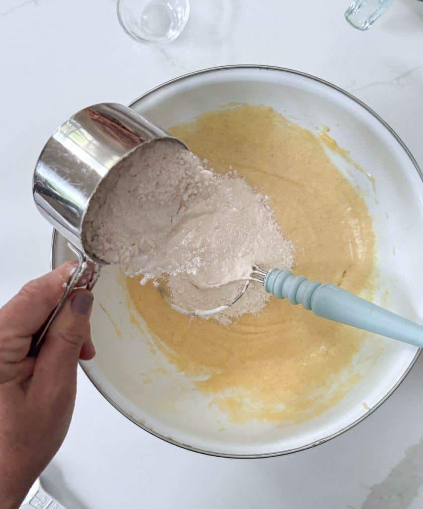 adding dry ingredients to the wet ingredients in a white bowl with a whisk in the bowl