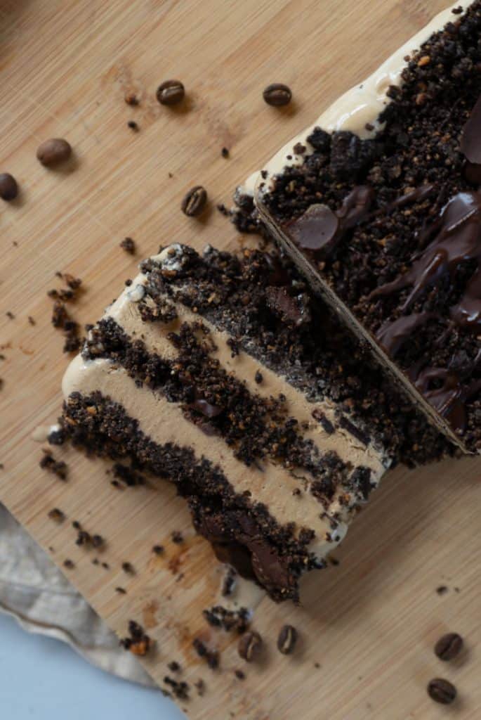 Coffee ice cream dessert on a wood plank with coffee beans strewn around and a slice of dessert laying next to the dessert