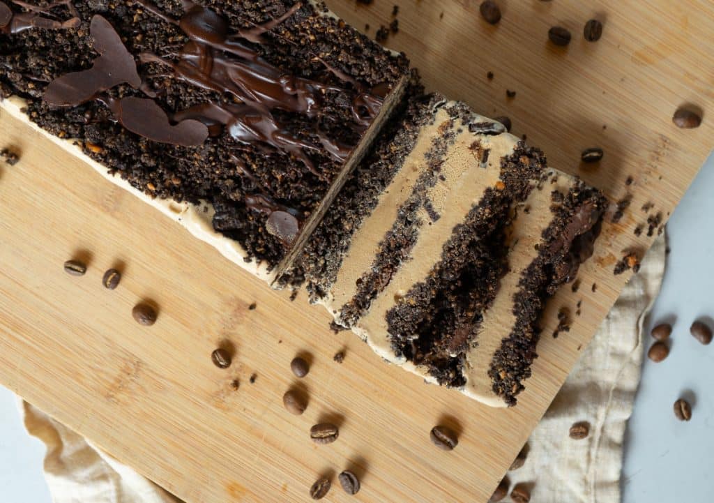 Coffee ice cream dessert on a wood plank with coffee beans strewn around and a slice of dessert laying next to the dessert