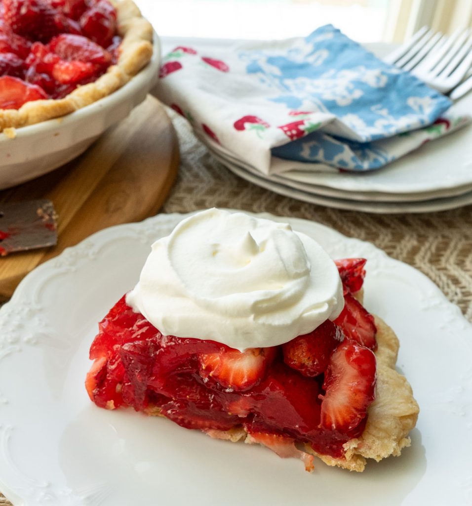 slice of fresh strawberry pie on a white plate with whole pie in the upper left corner plates, forks and napkins in upper right corner of the photo