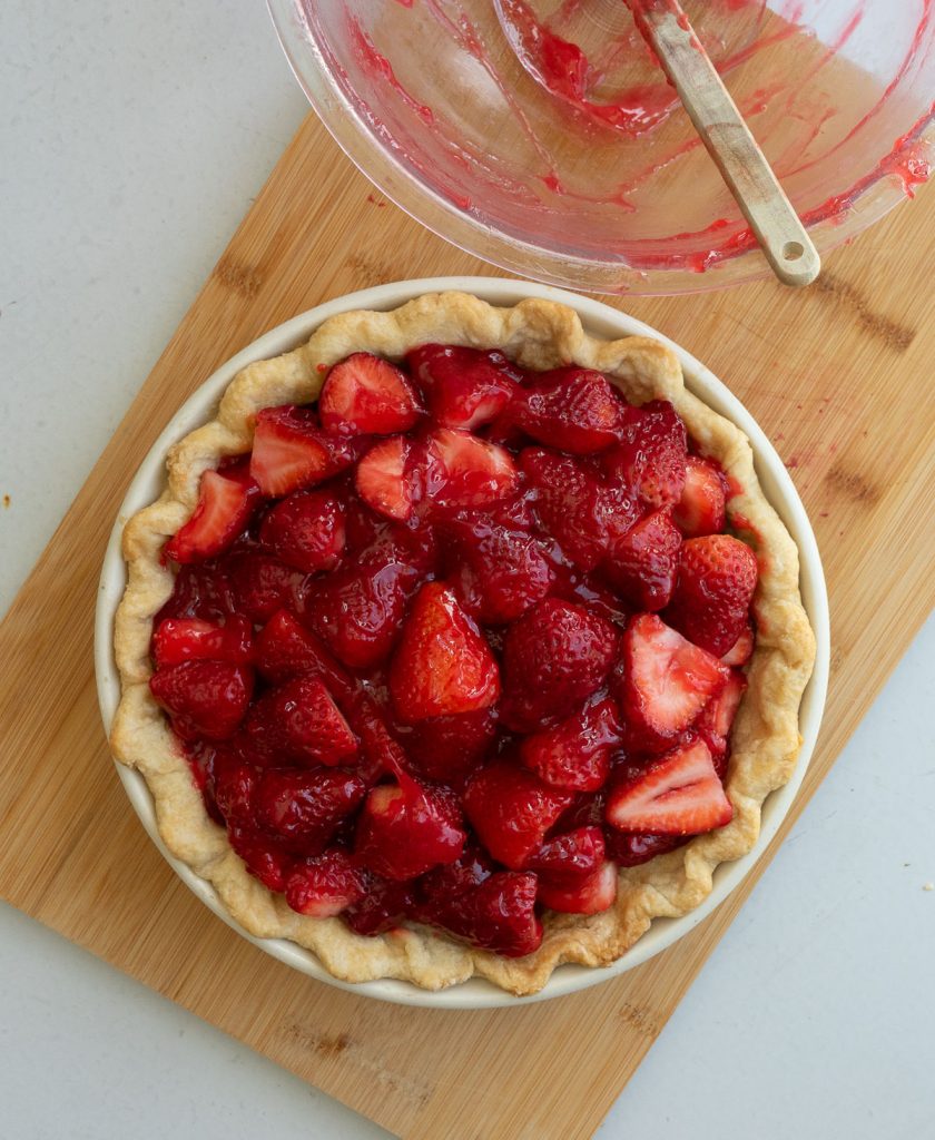fresh strawberry pie on a wood cutting board with empty glass bowl and a spatula to the top right of the pie