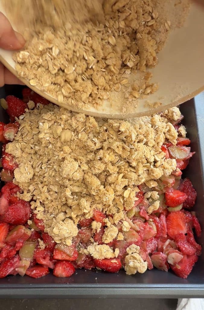 pouring crumble topping on top of fruit in a square baking dish