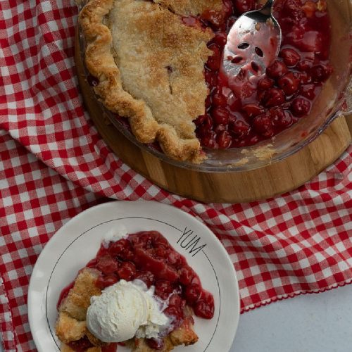 slice of cherry pie on a plate with a scoop of vanilla ice cream, whole pie is in the picture with a slice taken out