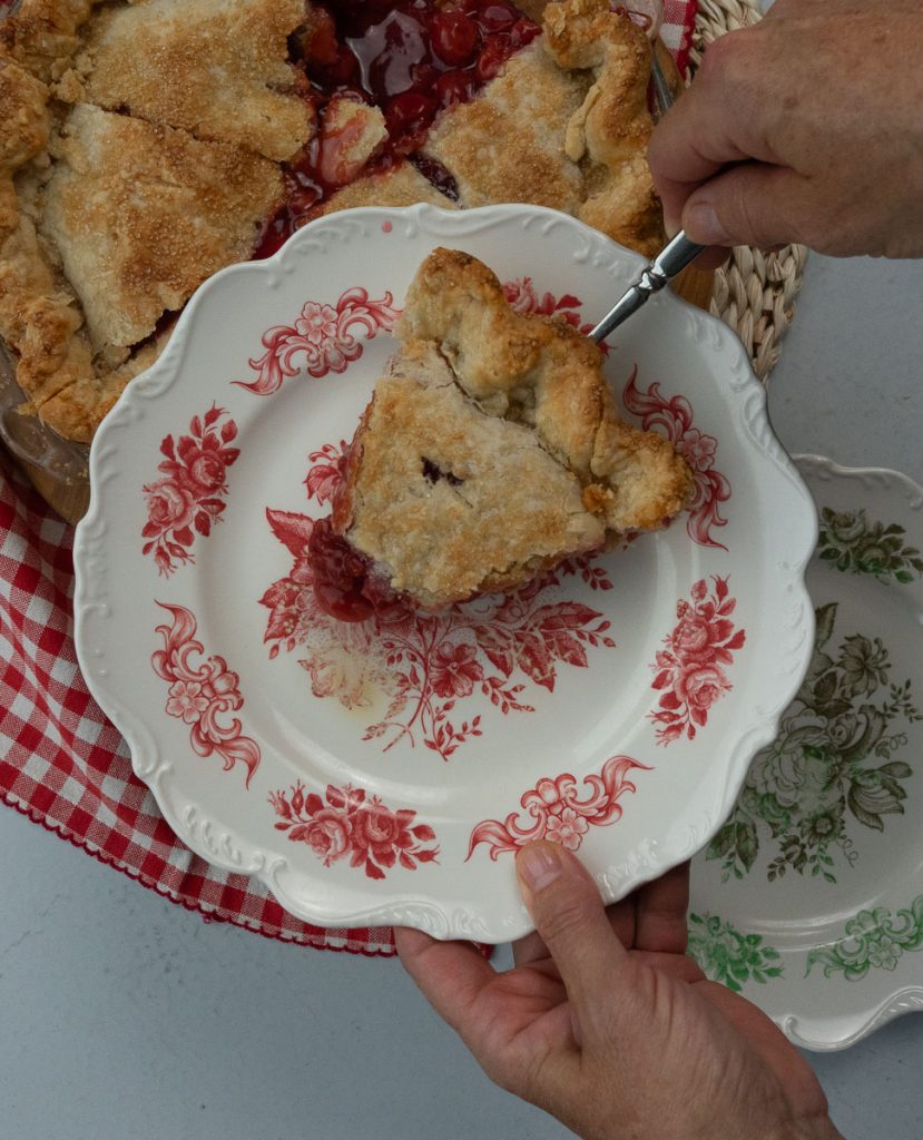 hands holding a plate with a slice of cherry pie whole cherry pie is in the background