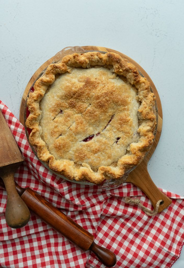 cherry pie sitting on a white counter top with rolling pin and red checked cloth in the bottom half of the photo