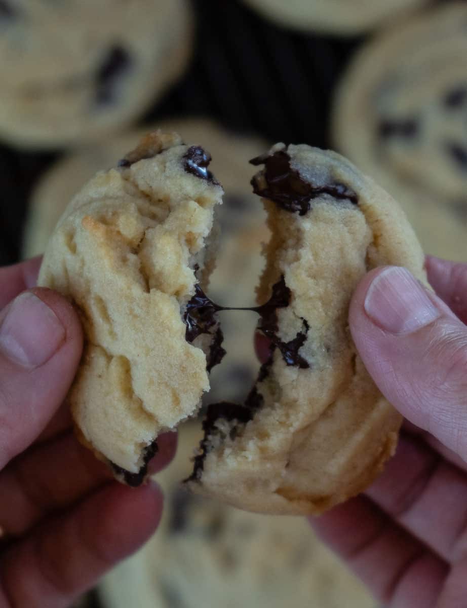 picture of hands cracking a warm chocolate chip cookie in half with the chocolate dripping out cookie is being held over a cooling rack of cookies