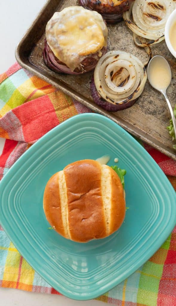 top view of burger on a teal fiestaware plate with hamburger toppings and condiments on a baking sheet