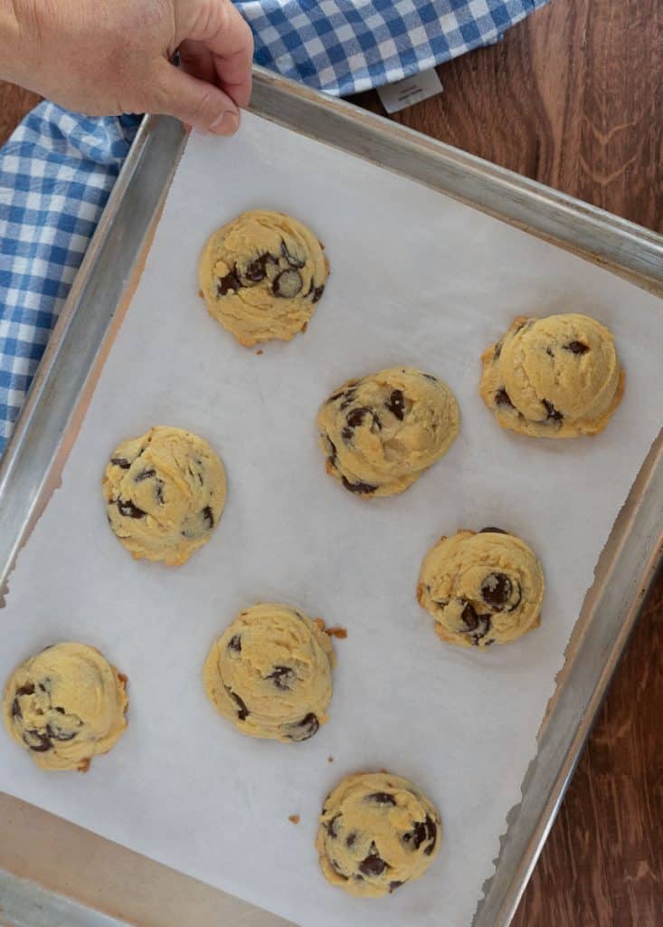 chocolate chip cookies on a parchment line baking sheet on a wood table with hands setting the sheet down a blue and white checked clothe is in the upper left hand corner of the photo