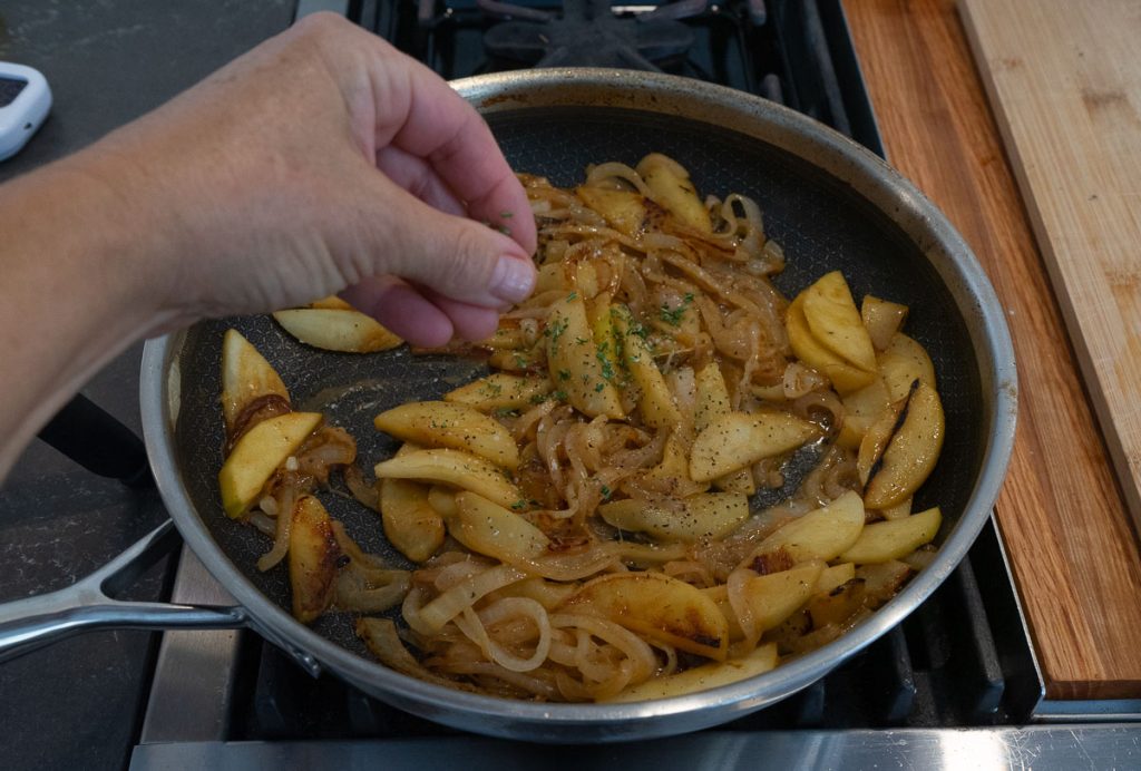 caramelized onions and apples in skillet on stovetop with a hand adding chopped rosemary