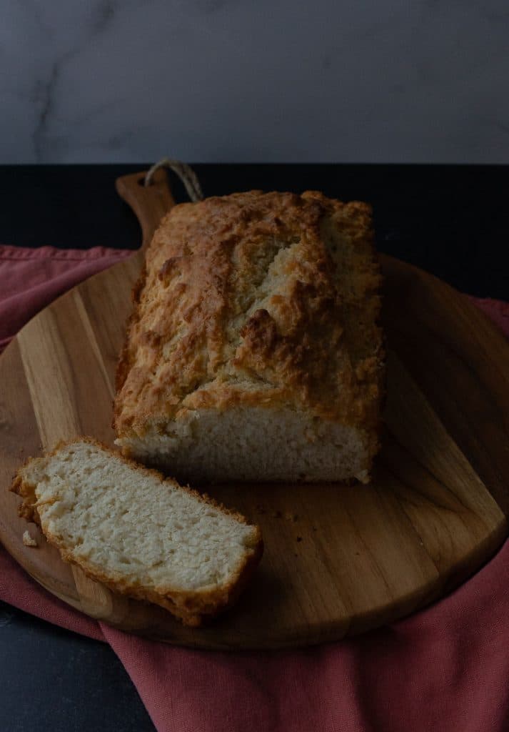 oaf of beer bread on a wood cutting board with a slice cut off and laying beside thee bread