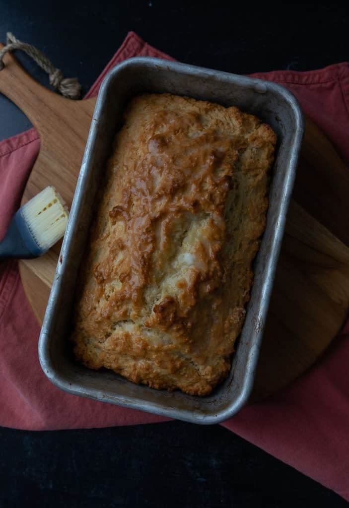 top view of a loaf of beer b read in a loaf pan sitting on a wood cutting board with the pastry brush laying on the left side