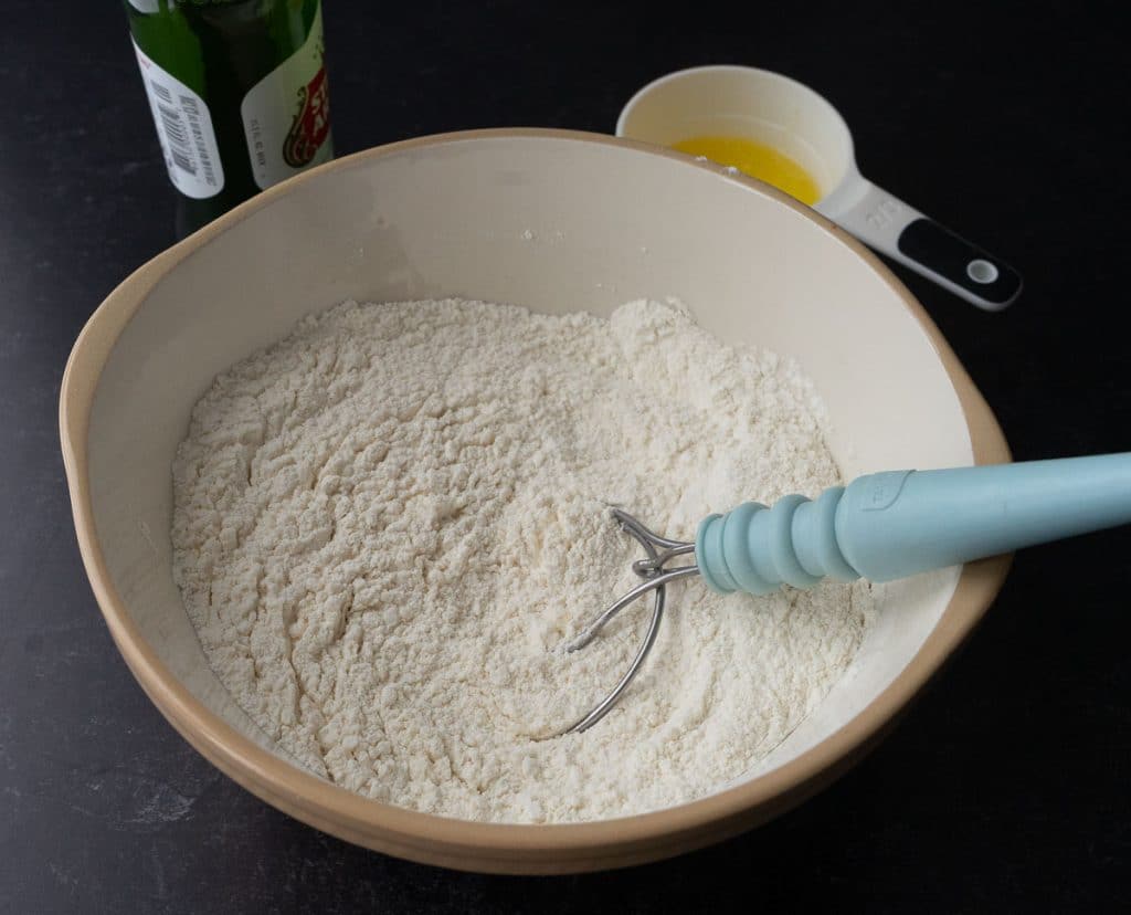 dry ingredients for beer bread mixed together in a glass bowl with a whisk in the bowl, a measuring cup of of melted butter and a beer are in the background