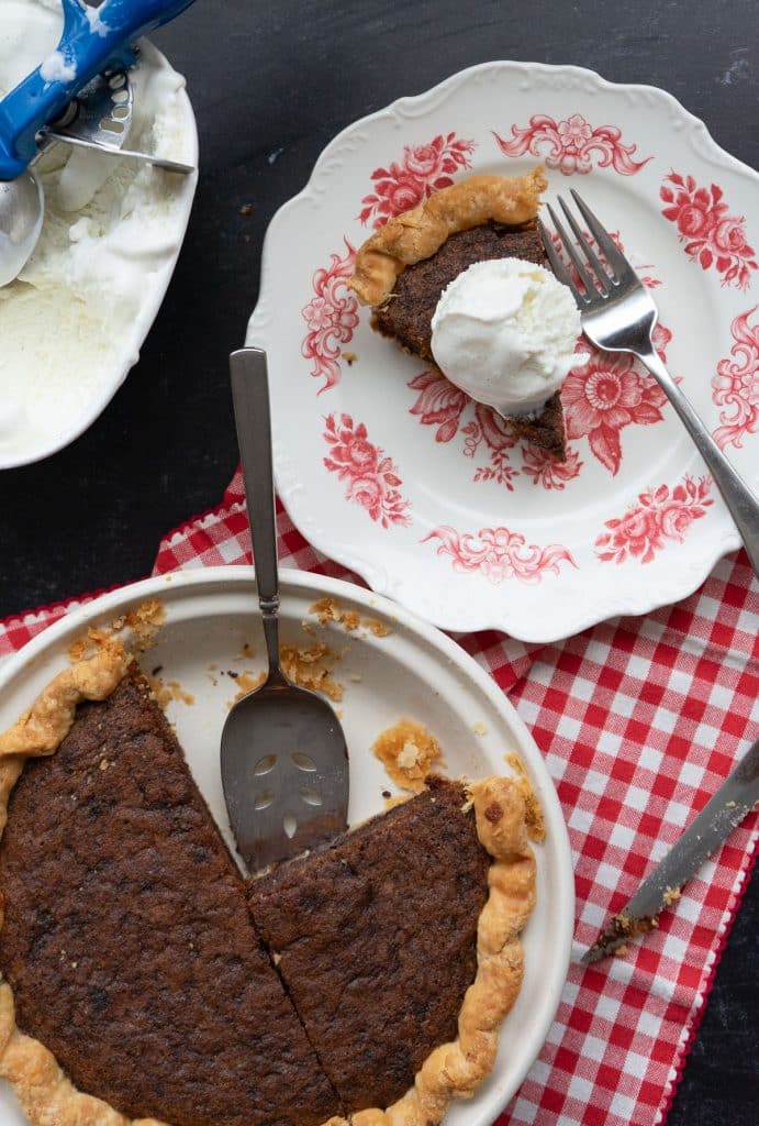 chocolate chip cookie pie with a piece removed a plate with a slice of pie and a scoop of ice cream on top a red gingham napkin is laying on the black countertop