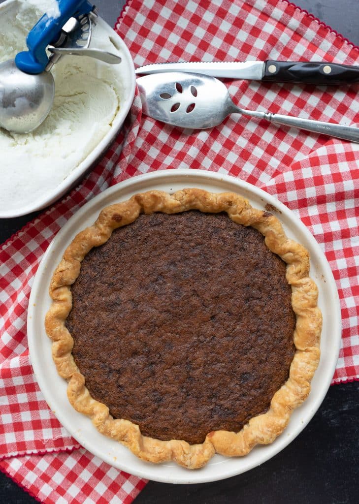whole toll house cookie pie with a knife, and a pie server laying above it and an open container of vanilla ice cream with a scoop in the upper left corner of the photo