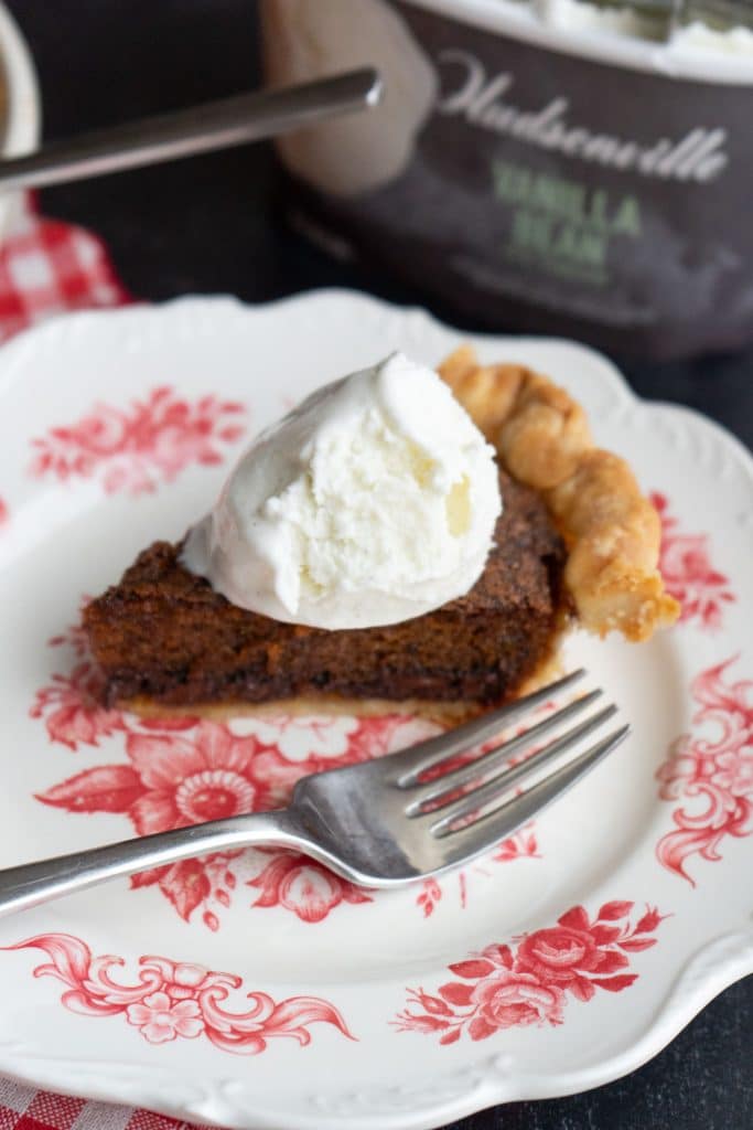 slice of toll house chocolate chip cookie pie on a plate with a fork setting beside it on the plate there is ice cream on pie and a container of vanilla ice cream in the background