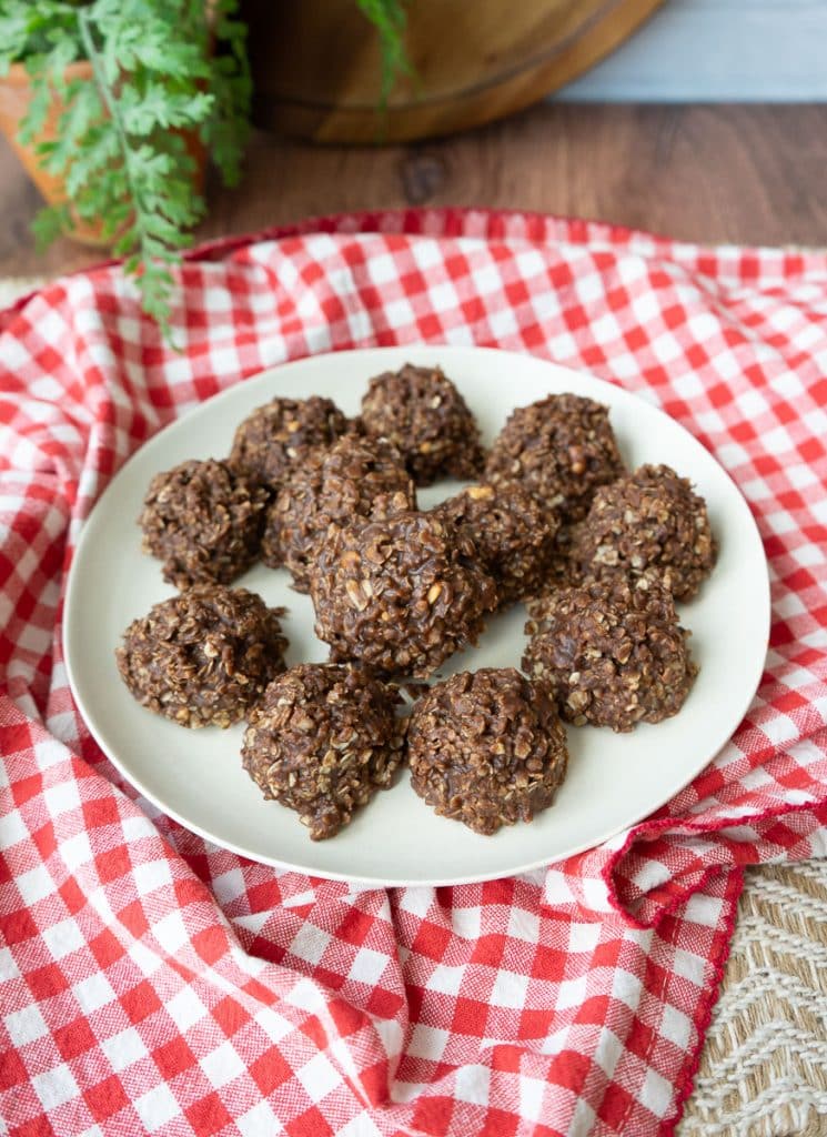 plate of no-bake cookies on a red and white checked cloth there is a wood cutting board and a plant in the background