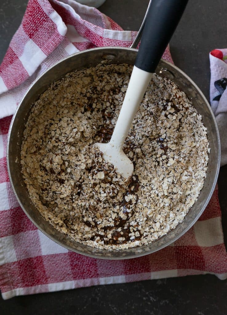 adding oatmeal to the chocolate peanut butter mixture in the saucepan there is a white and black spatula in the pan pan is sitting on a red and white kitchen towel