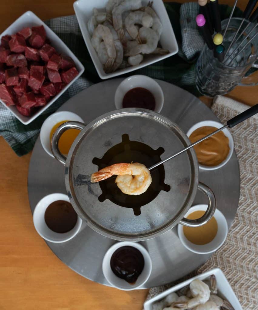 shrimp on a fondue fork over a fondue pot with sauces and proteins around the fondue pot