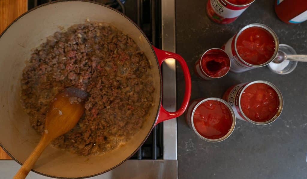 browning ground beef in dutch oven on the stove top with the cans of opened tomato products open next to the stovetop