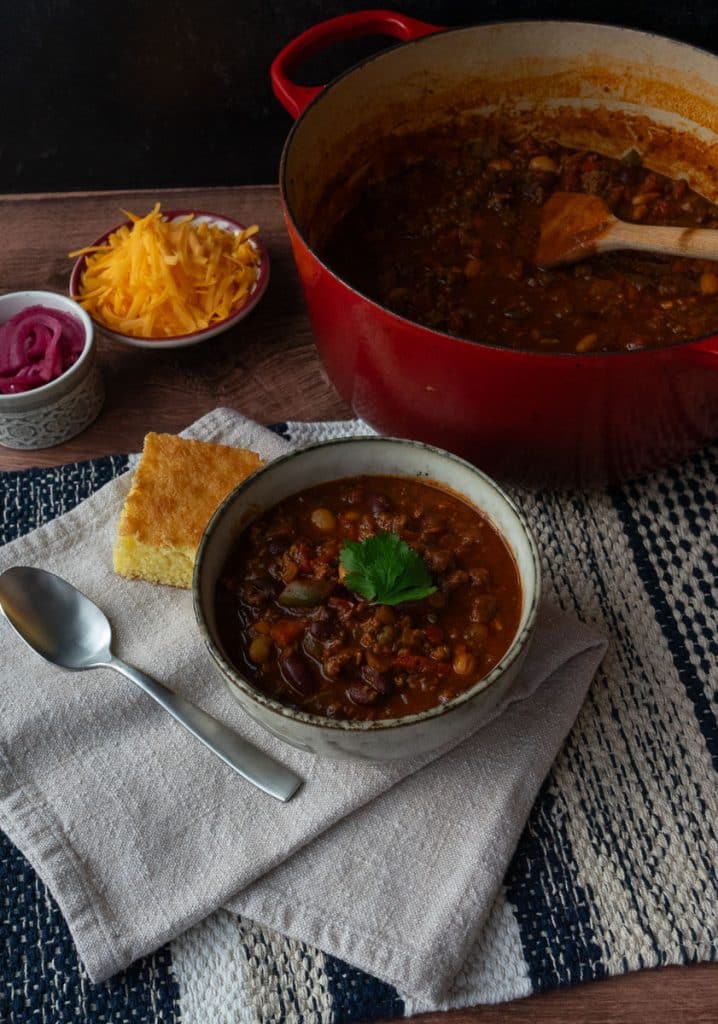 bowl of chili with the pot of chili to the rt and corn bread on the side with a bowl of chedda and a bowl of pico de gallo in back ground