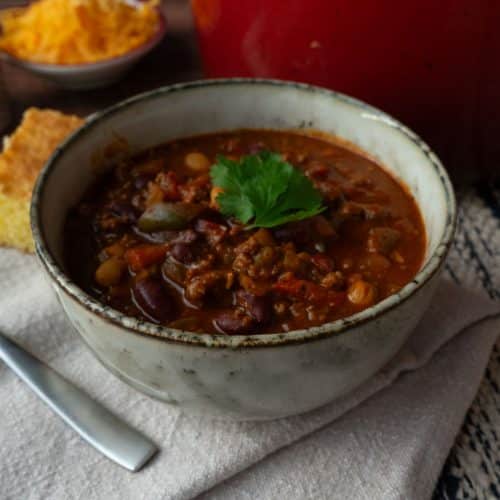bowl of chili with the pot of chili to the rt and corn bread on the side with a bowl of chedda and a bowl of pico de gallo in back ground