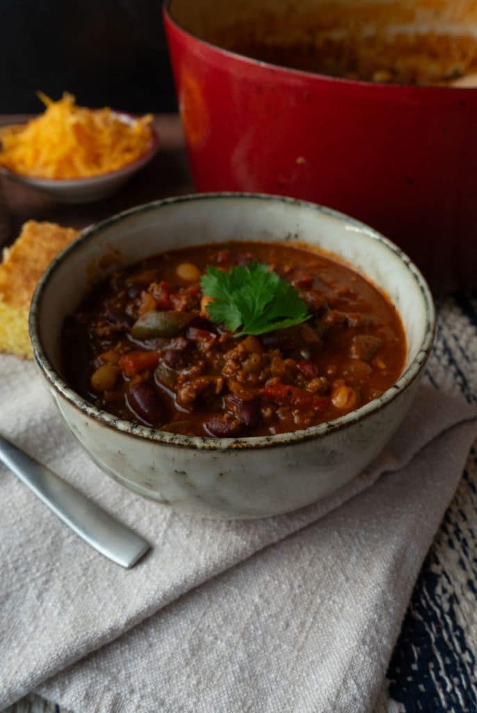 bowl of chili with the pot of chili to the rt and corn bread on the side with a bowl of chedda and a bowl of pico de gallo in back ground