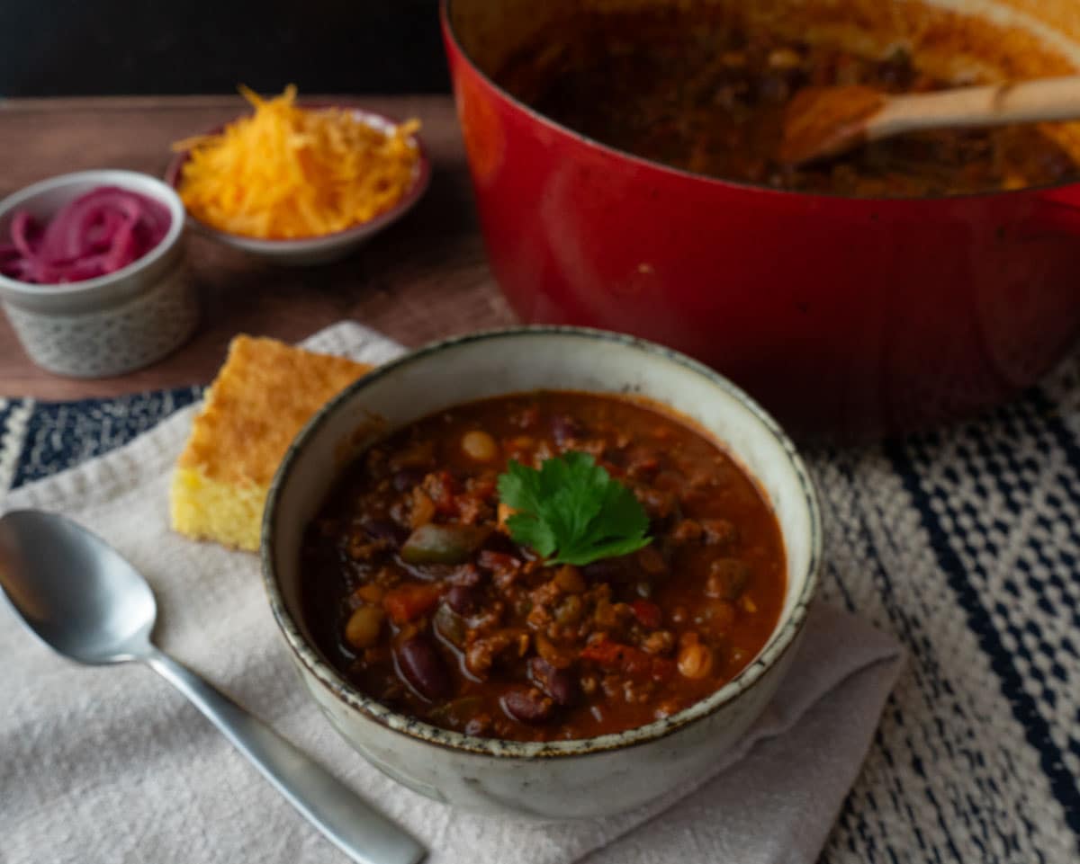 bowl of chili with the pot of chili to the rt and corn bread on the side with a bowl of chedda and a bowl of pico de gallo in back ground