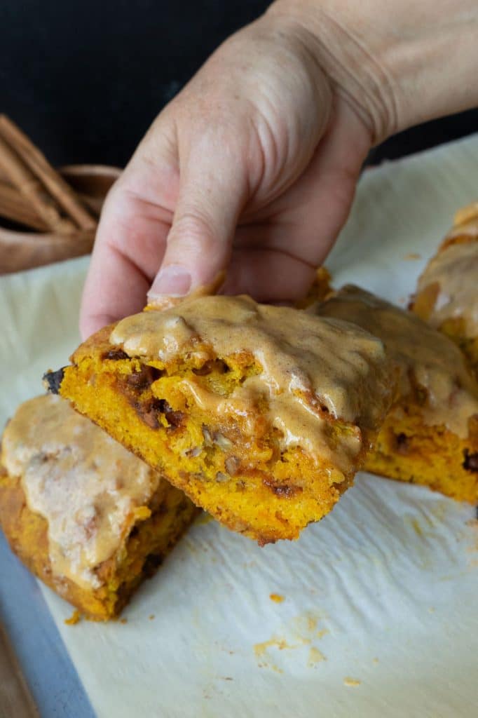 hand holding a pumpkin scone over the baking sheet lined with parchment paper with scones on it. in background there is a small wood bowl filled with cinnamon sticks