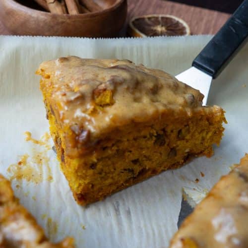 pumpkin scones sitting on a parchment lined baking sheet. there is one scone that is the center or the photo behind is a black backsplash and a wood cup with cinnamon sticks in it
