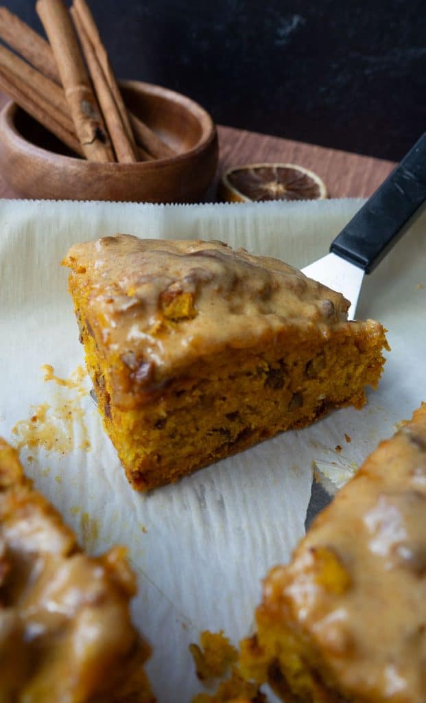 pumpkin scones sitting on a parchment lined baking sheet. there is one scone that is the center or the photo behind is a black backsplash and a wood cup with cinnamon sticks in it