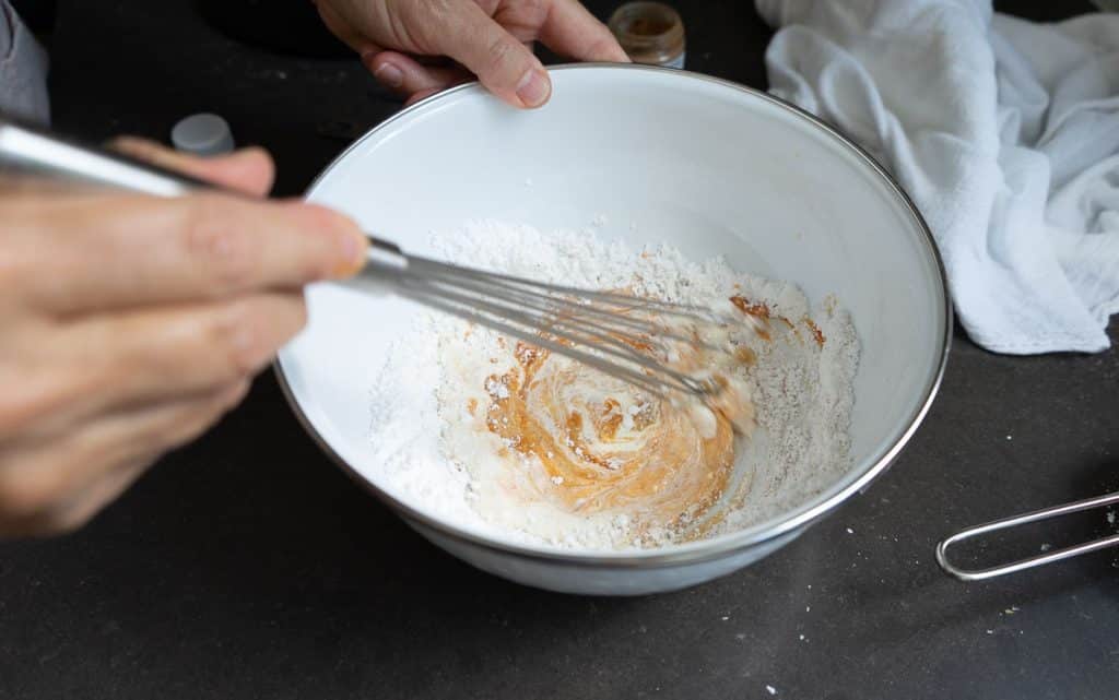 whisking together the pumpkin spiced glaze in a white bowl with a straight whisk. bowl is setting on a black countertop. there is a white kitchen towel in the upper right hand corner of the picture