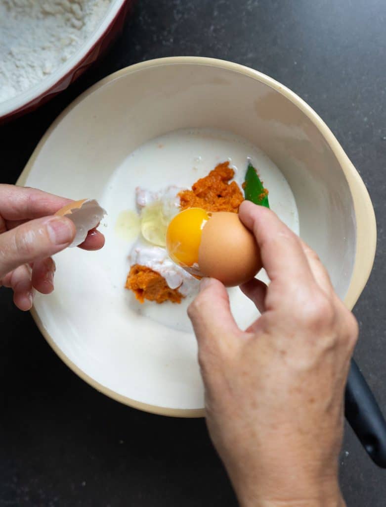 mixing wet ingredients in a glass bowl. hand is cracking egg into buttermilk and pumpkin puree already in bowl