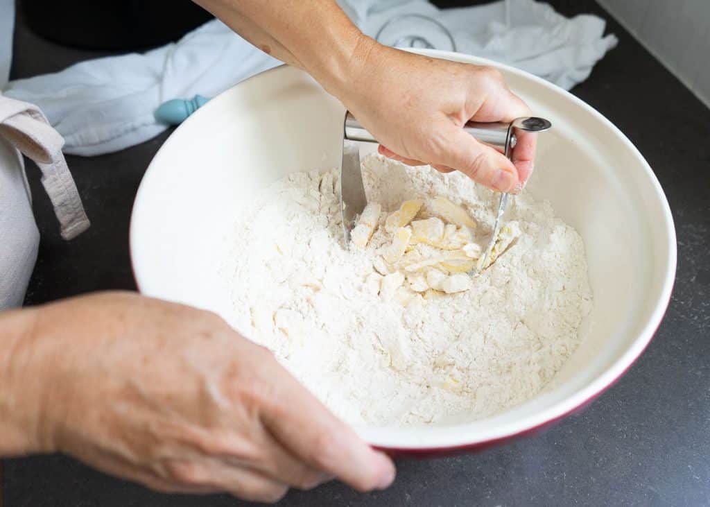 cutting butter into dry ingredients in white mixing bowl. one hand is using a pastry cutter the other hand is holding the bowl. bowl is on a black countertop there is a kitchen towel in the background