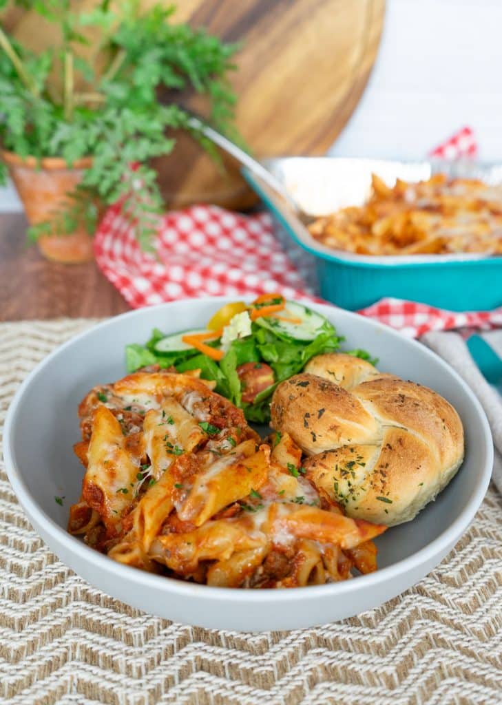 mostaccioli plated on a gray plate on a tan placemat with a side salad and a garlic roll the casserole dish of mostaccioli is behind the dish sitting on a red and white checked cloth there is a plant in the upper left corner all is setting on a wood table
