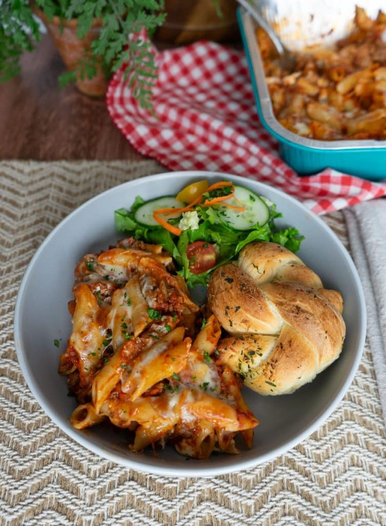 mostaccioli plated on a gray plate on a tan placemat with a side salad and a garlic roll the casserole dish of mostaccioli is behind the dish sitting on a red and white checked cloth there is a plant in the upper left corner all is setting on a wood table
