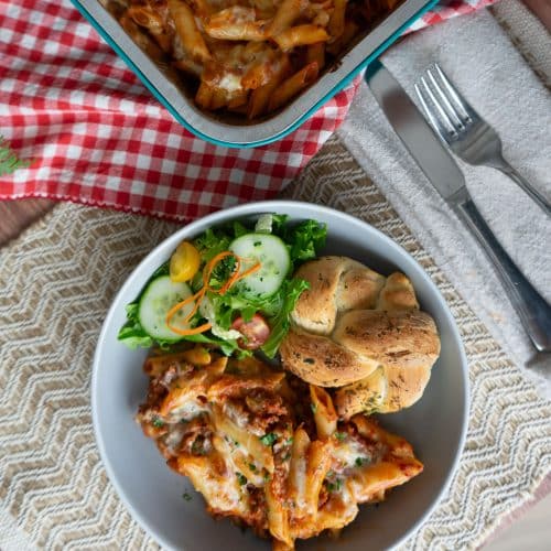 top view of plated mostaccioli and a green salad on the side with a garlic roll a corner of the casserole dish is visible in the upper right corner