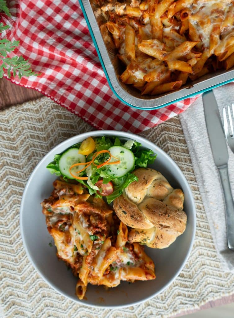 top view of plated mostaccioli and a green salad on the side with a garlic roll a corner of the casserole dish is visible in the upper right corner