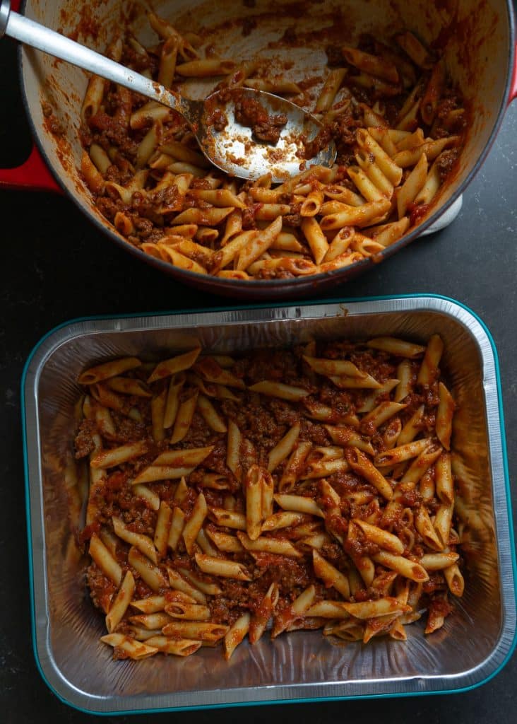 pot of noodles and sauce, with a 9x13 pan sitting in from of the pot, half of the noodles and sauce have been placed in the bottom of the baking dish