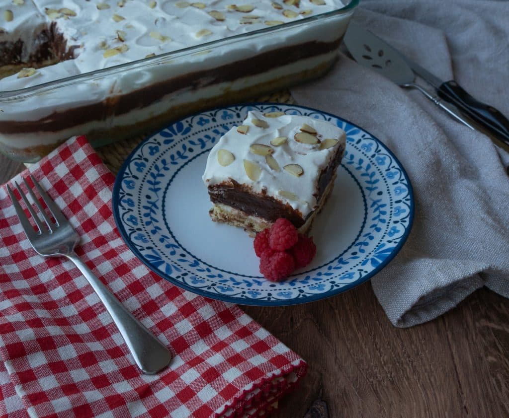 serving of dessert on a plate with a red checked napkin the left with a fork and the dish of dessert with one square cut out in the background