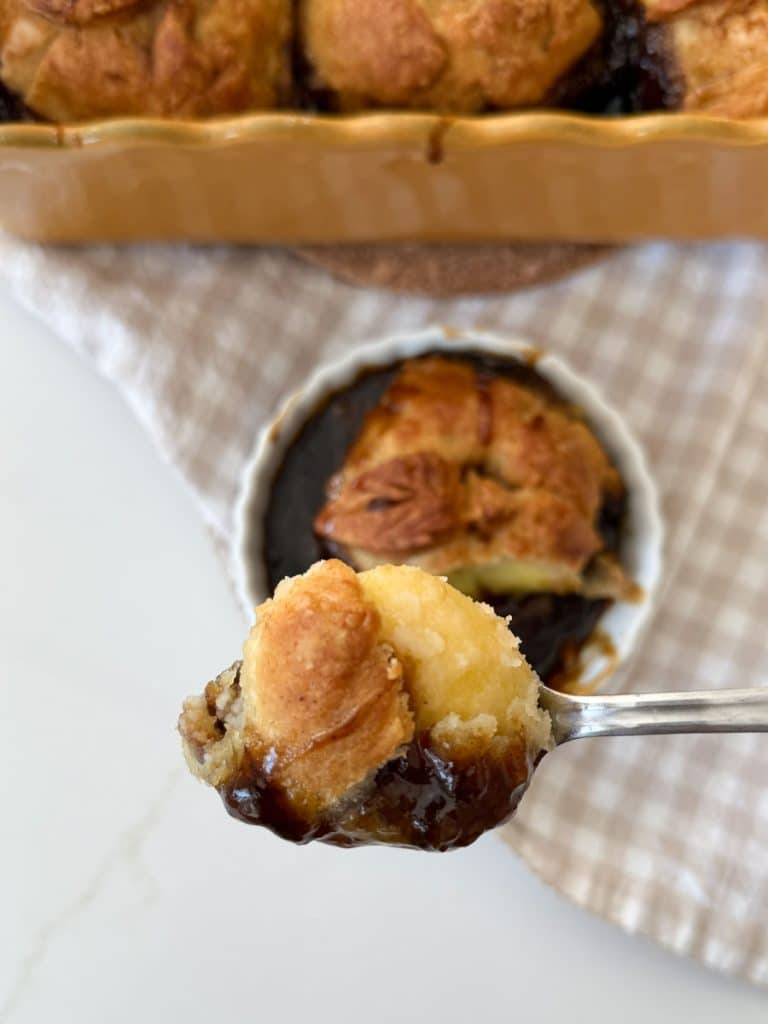 a bite of the apple dumpling on a spoon hovering over the an apple dumpling in a white dish with the pan of apple dumplings in the background