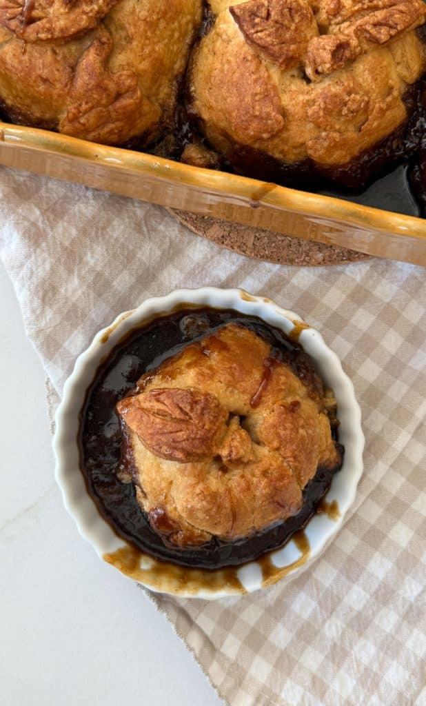 an apple dumpling in a white dish with the pan of apple dumplings in the background