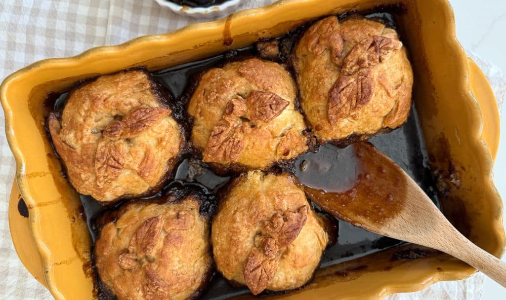 apple dumplings in a baking dish with one missing and a wooden spoon in the dish where the apple dumplings was dish is sitting on a tan checked cloth