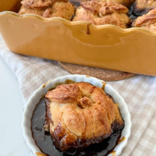 an apple dumpling in a white dish with the pan of apple dumplings in the background