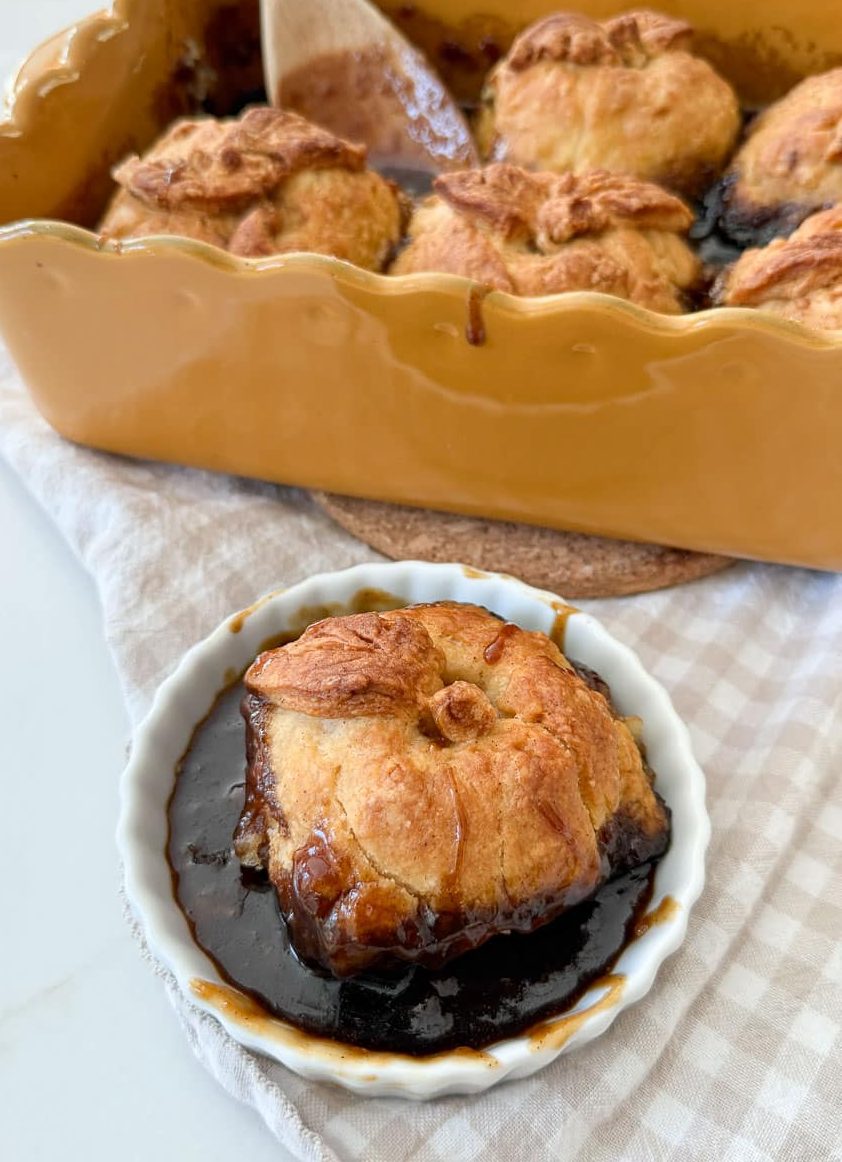 an apple dumpling in a white dish with the pan of apple dumplings in the background