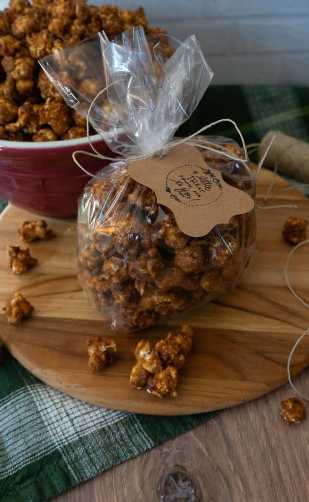bag of caramel corn with a gift tag on it, sitting beside a bowl of caramel corn on a wood cutting board